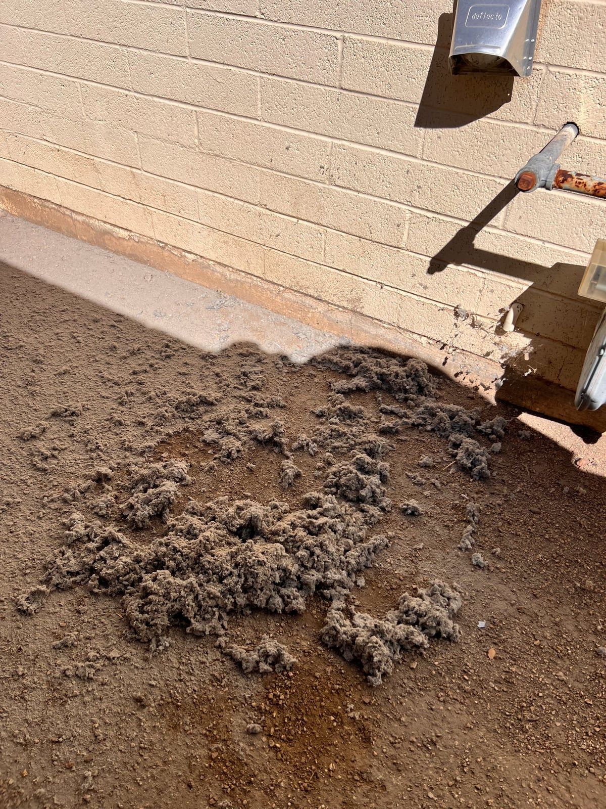 A pile of gray dryer lint on the dirt below an exterior dryer vent hood on a block wall