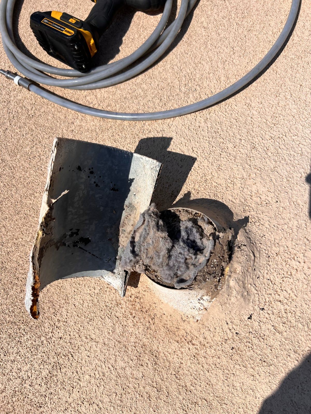 A dryer vent opening on a flat roof packed with gray lint, its rusted cap lying beside it and a cleaning rod coiled nearby